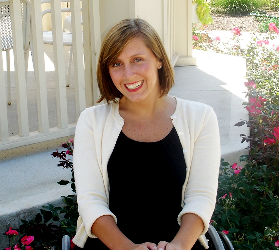 Amber Ohaver smiles in front of a flower garden. She's wearing a cardigan and sits in a manual wheelchair.